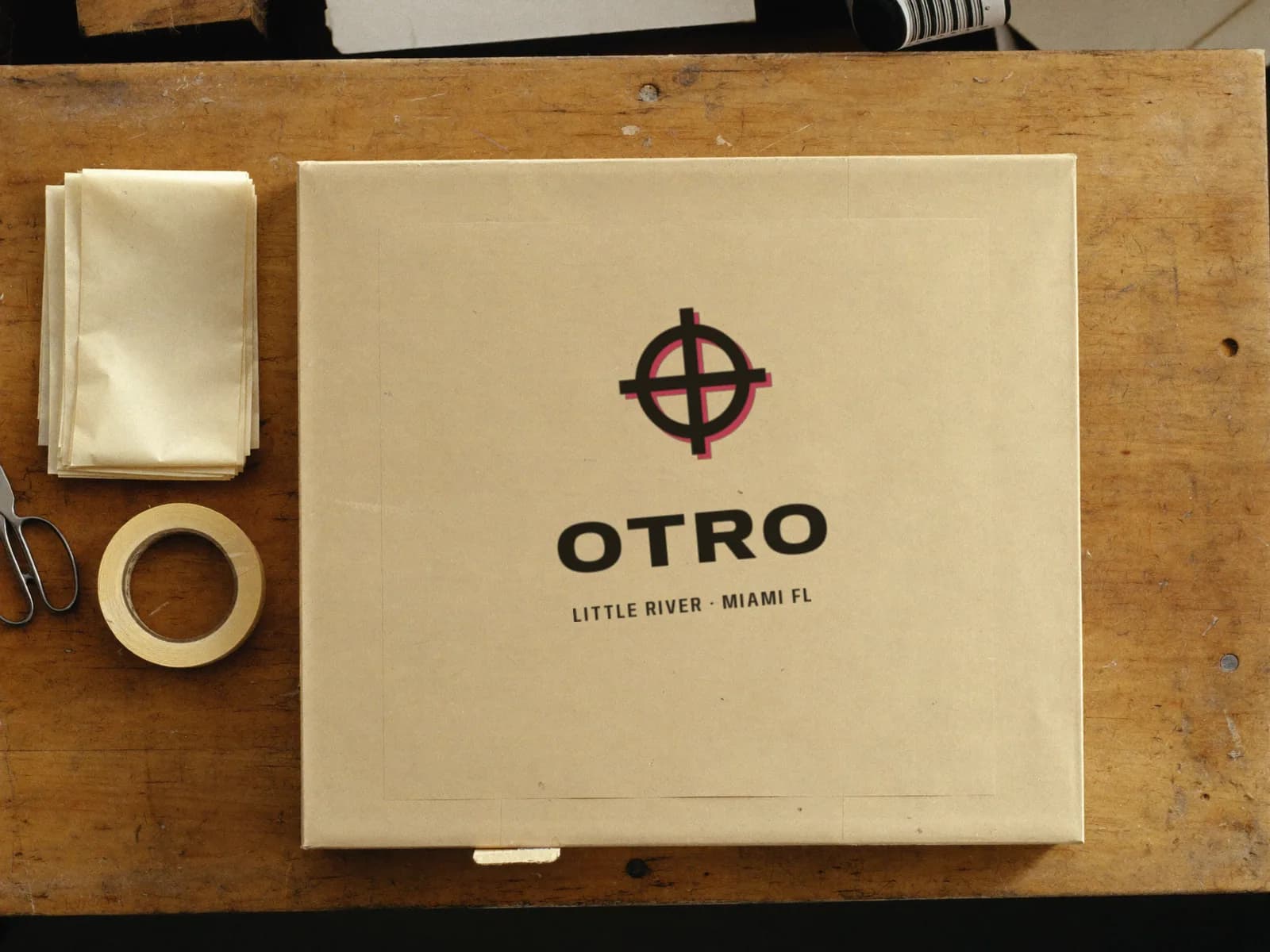 A plain kraft mailer box on a plywood packing table, stamped with the OTRO lockup
